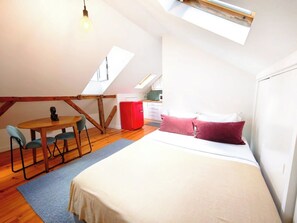Una habitación compacta con cama, una pequeña mesa y sillas, un electrodoméstico rojo y un tragaluz.