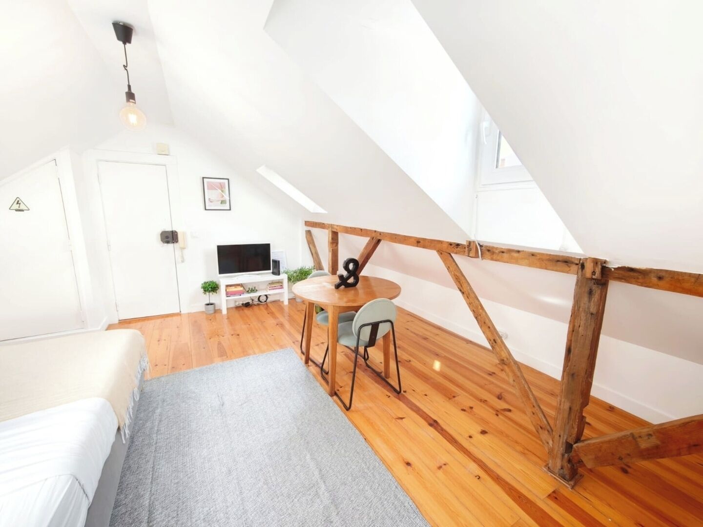 A loft bedroom with a sloped ceiling, a bed, a small table with chairs, a television, and a wooden beam structure.