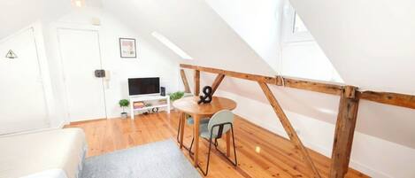 A loft bedroom with a sloped ceiling, a bed, a small table with chairs, a television, and a wooden beam structure.