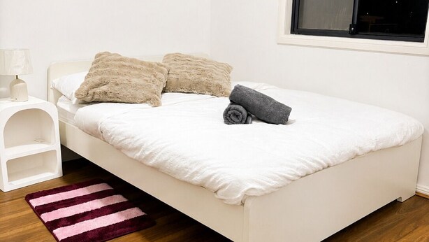 Een eenpersoonskamer met een wit bed, nachtkastje, lamp, raam en een houten vloer.