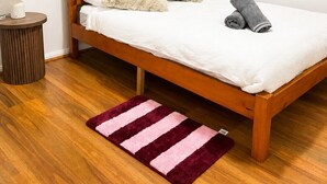 Een enkel bed met houten frame, wit beddengoed, twee kussens en een opgerolde handdoek. Er staat een klein nachtkastje met een lamp en een rond vloerkleed met roze strepen.