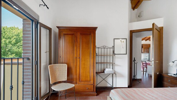 Una camera da letto con un letto, un'armadiatura in legno, una sedia in vimini, un'unità scaffale in metallo e vista sull'esterno tramite una porta scorrevole.