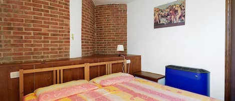 Ein Zimmer mit Backsteinwand, einem Bett mit gemustertem Überwurf, einem kleinen Tisch mit Lampe und einem Mülleimer.