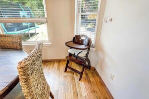 A room with a wooden floor, a wicker chair, a small table with a chair, and a window with blinds. - Suncrest Rise! Family Escape Near Fort Carson (Colorado Springs)