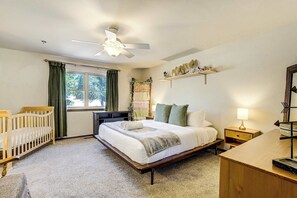 A bedroom with a bed, two cribs, a ceiling fan, a window with curtains, a shelf with decorative items, and a nightstand with a lamp. - Suncrest Rise! Family Escape Near Fort Carson (Colorado Springs)