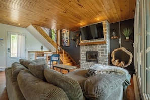 A cozy living room with a wooden ceiling, a stone fireplace, a television mounted above it, and a staircase leading to an upper level. - La paix dans le Nord | Lac Brulé | SPA | SAUNA (Matawinie Regional County Municipality)