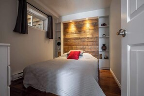 A bedroom with a bed, a wooden accent wall, a window with curtains, and a shelf with decorative items. - La paix dans le Nord | Lac Brulé | SPA | SAUNA (Matawinie Regional County Municipality)