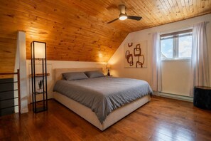 A bedroom with a sloped ceiling, a bed, a standing lamp, a window with curtains, and a ceiling fan. - La paix dans le Nord | Lac Brulé | SPA | SAUNA (Matawinie Regional County Municipality)