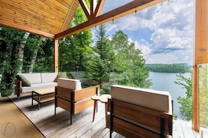 An outdoor deck with wooden furniture, a glass balustrade, and a view of a lake surrounded by trees. - La paix dans le Nord | Lac Brulé | SPA | SAUNA (Matawinie Regional County Municipality)