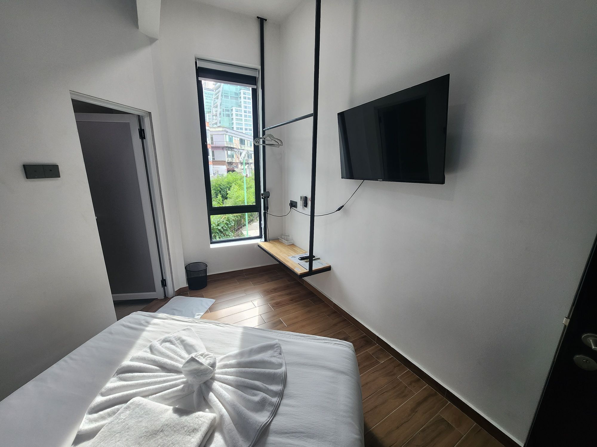 A hotel room with a bed, a TV mounted on the wall, a window with a view of buildings, and a door.