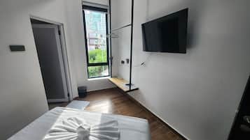 A hotel room with a bed, a TV mounted on the wall, a window with a view of buildings, and a door.