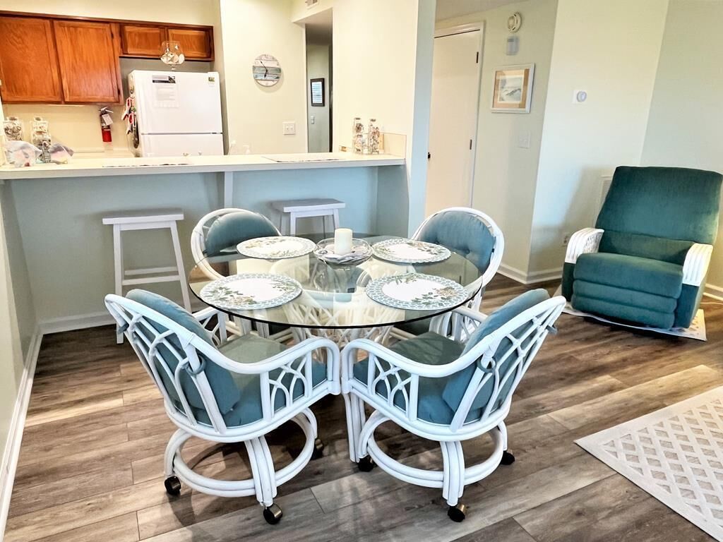 Un comedor con una mesa redonda de vidrio rodeada por cuatro sillas blancas con cojines azules, una zona de cocina con armarios de madera y un refrigerador blanco, y un sillón azul turquesa en la esquina.