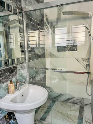 A bathroom with a glass-enclosed shower, a white sink, and marble tiles. - The Bella Villas Luxury Apartments (Lusaka)
