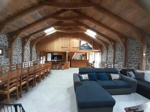 A spacious living area with a high ceiling, wooden beams, stone walls, a large sofa, and a kitchen in the background.