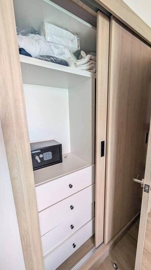 A wardrobe with a combination of drawers and hanging space, a small safe, and a door with a handle. - Cana Life Luxury | City Beach Pool View Suite (Punta Cana)