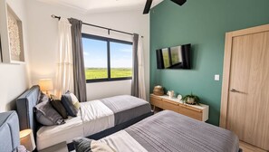A bedroom with two beds, a TV on the wall, a window with curtains, a bedside table with a lamp, and a view of the outdoors. - Cana Life Luxury | City Beach Pool View Suite (Punta Cana)