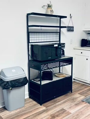 A kitchen area with a black metal shelf unit, a trash bin, and a coffee maker.