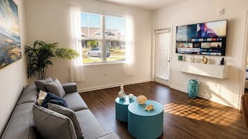A living room with a sofa, two round coffee tables, a ceiling fan, a TV mounted on the wall, and a window with blinds.