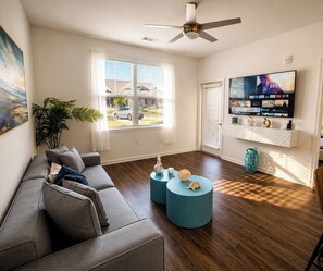 A living room with a sofa, two round coffee tables, a ceiling fan, a TV mounted on the wall, and a window with blinds. - Oasis by the Sea (Jacksonville)