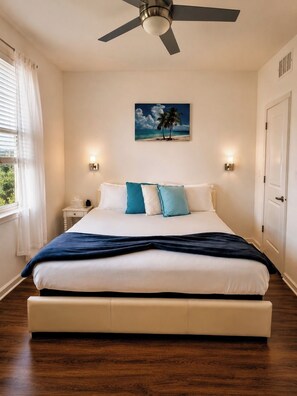 A bedroom with a bed, a ceiling fan, a painting of a beach scene, and a nightstand with a lamp. - Oasis by the Sea (Jacksonville)
