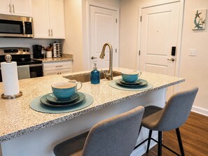 A kitchen with a central island, two bar stools, a microwave, a coffee maker, and a sink. - Oasis by the Sea (Jacksonville)