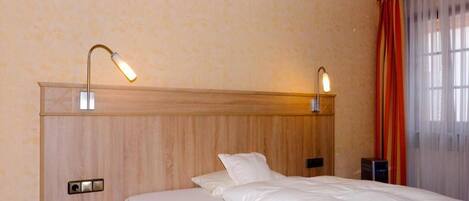 Habitación de hotel con cabecera de madera, cama con ropa blanca, dos lámparas de noche, teléfono en la mesita y una ventana con cortinas.