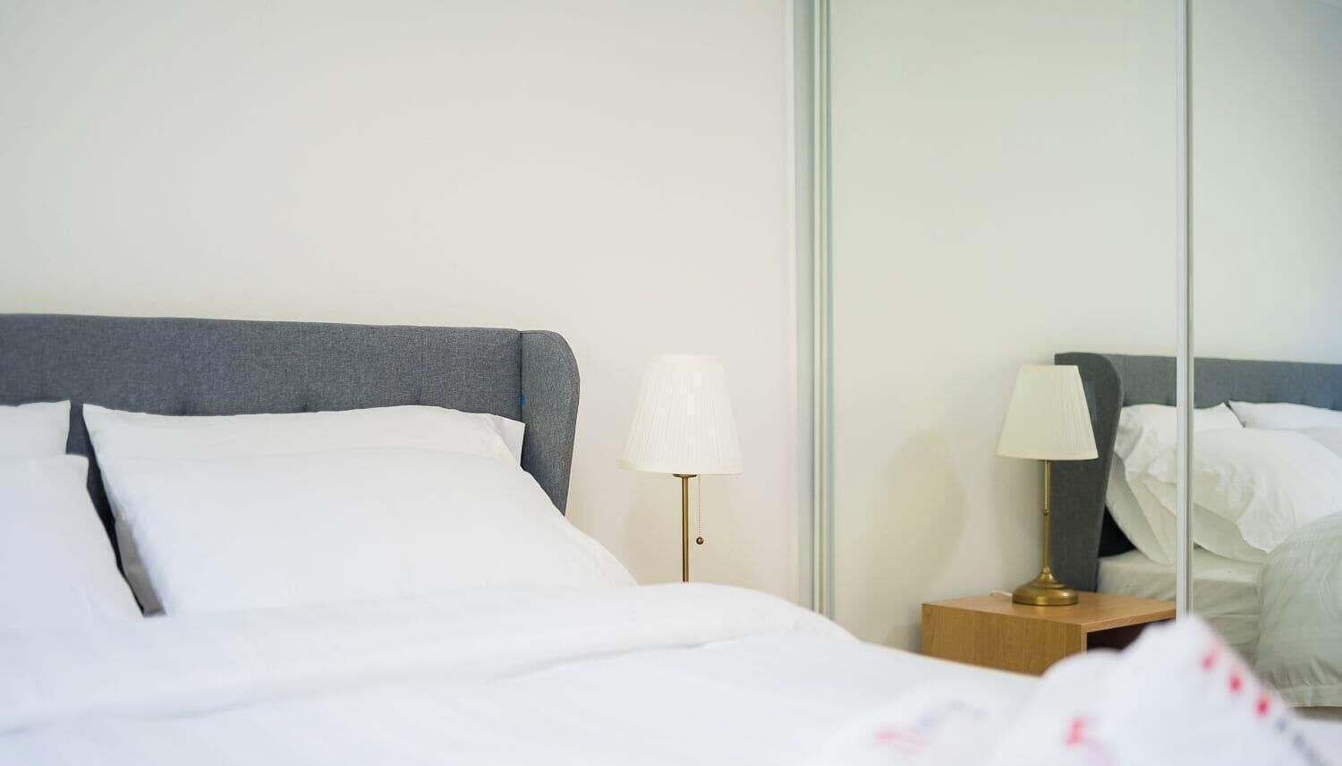 A neatly made bed with white bedding and a grey headboard, accompanied by a bedside table with a lamp.