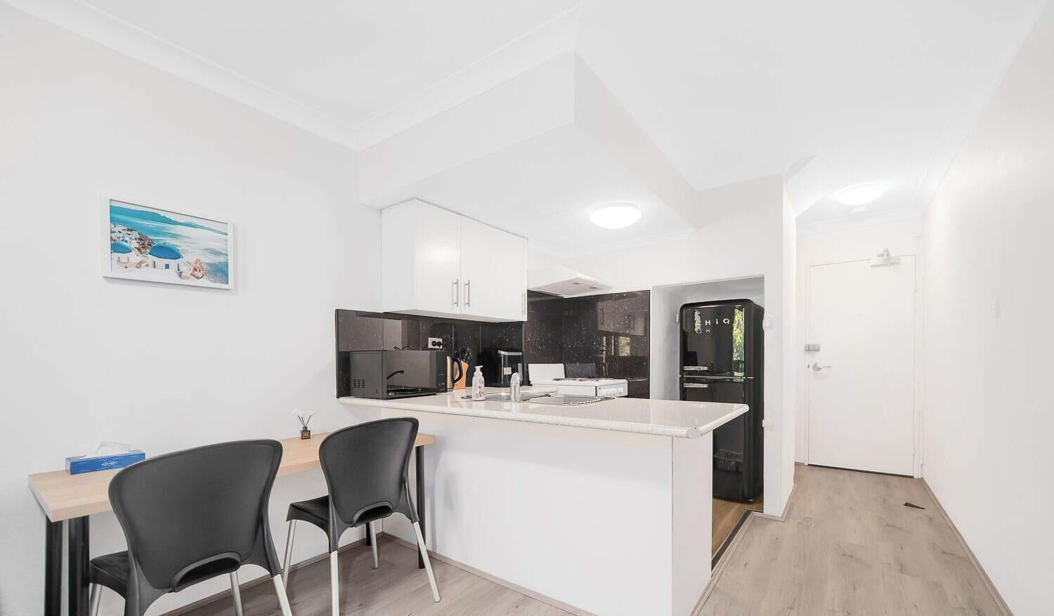 Una cocina compacta con armarios blancos, un refrigerador negro y un pequeño comedor con dos sillas.