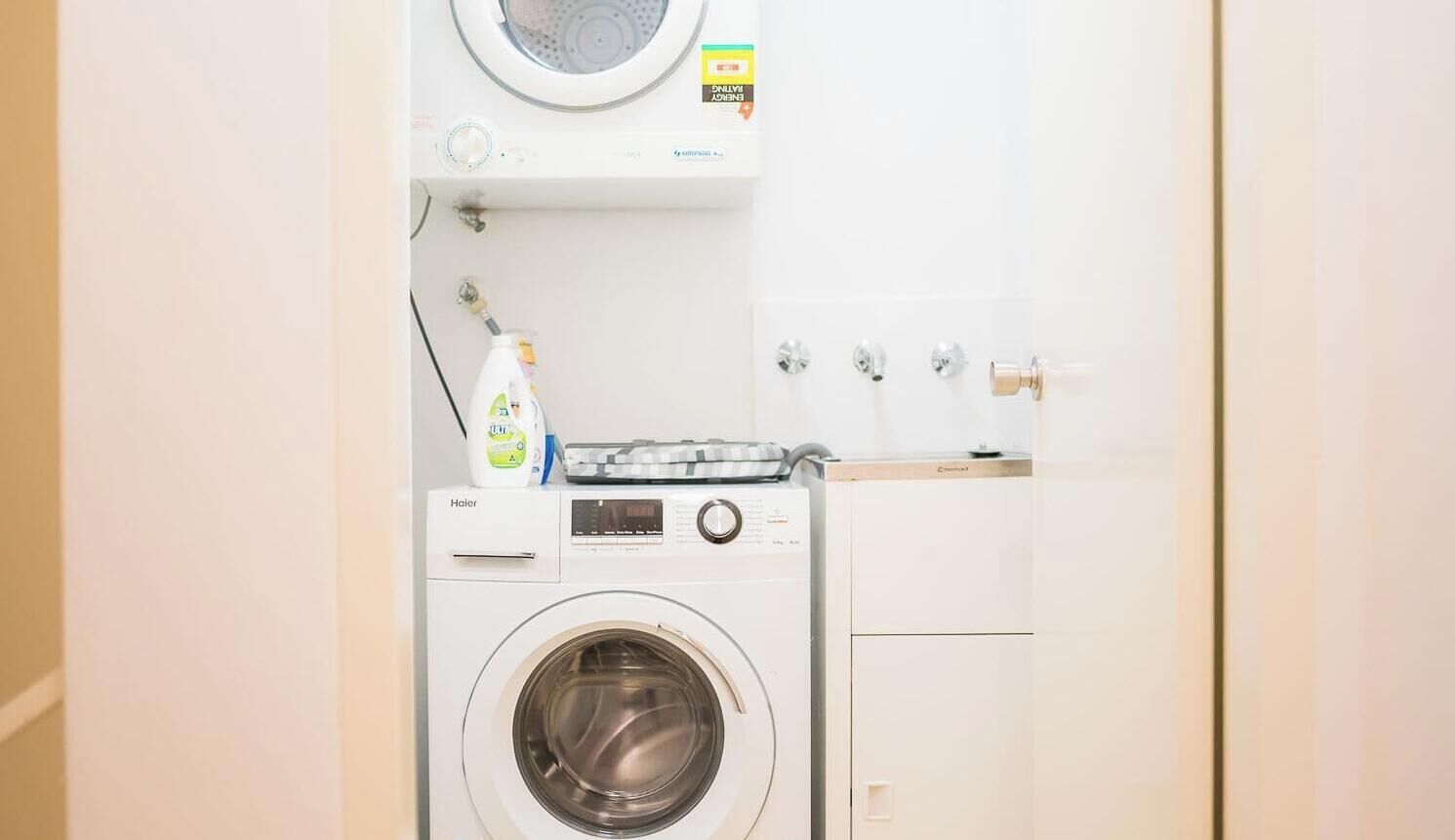 A compact, white laundry room with a front-loading washing machine, a drying rack, and a small cabinet.