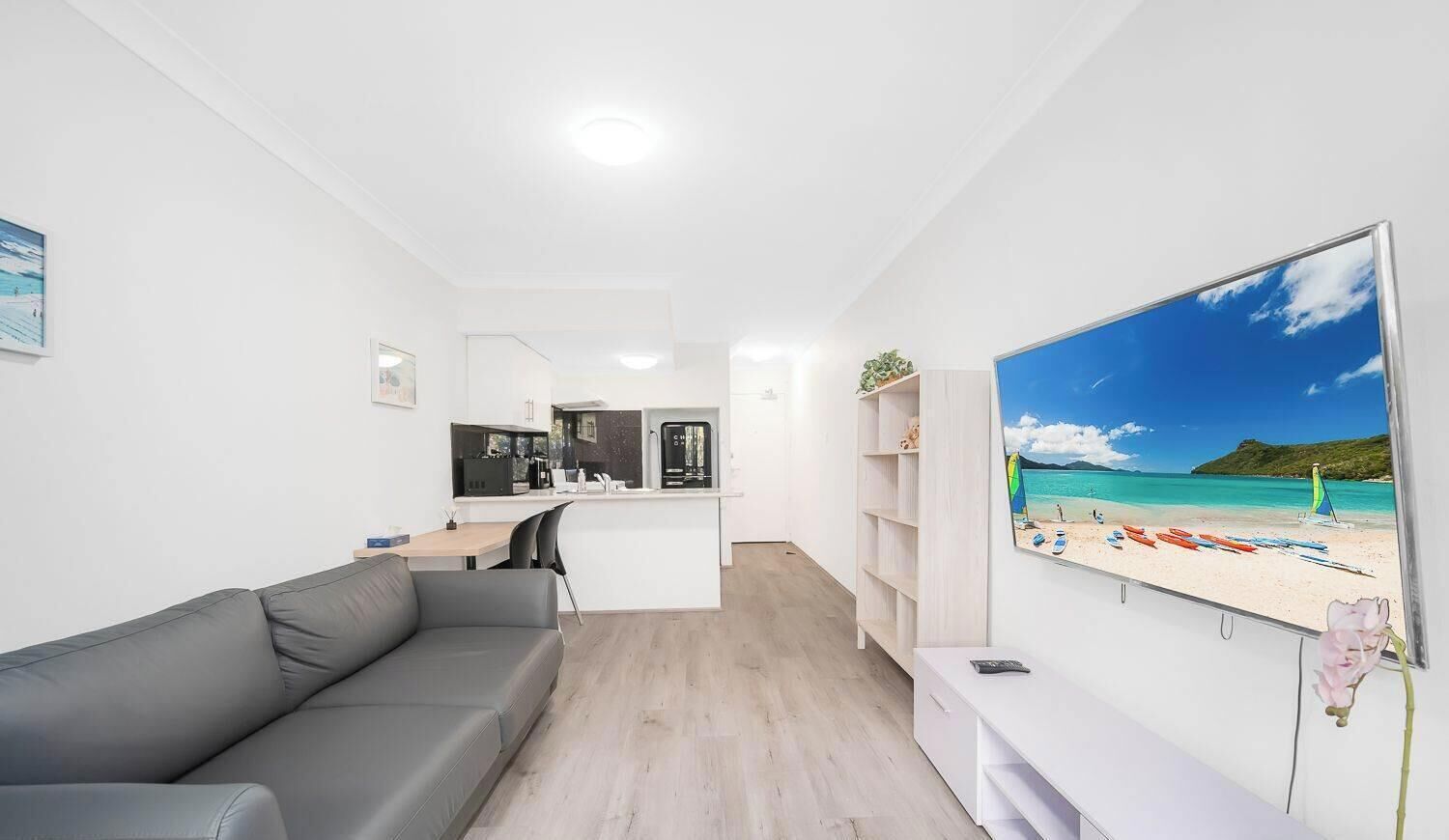 A modern living space with a grey sofa, a kitchen area, and a large TV displaying a beach scene.