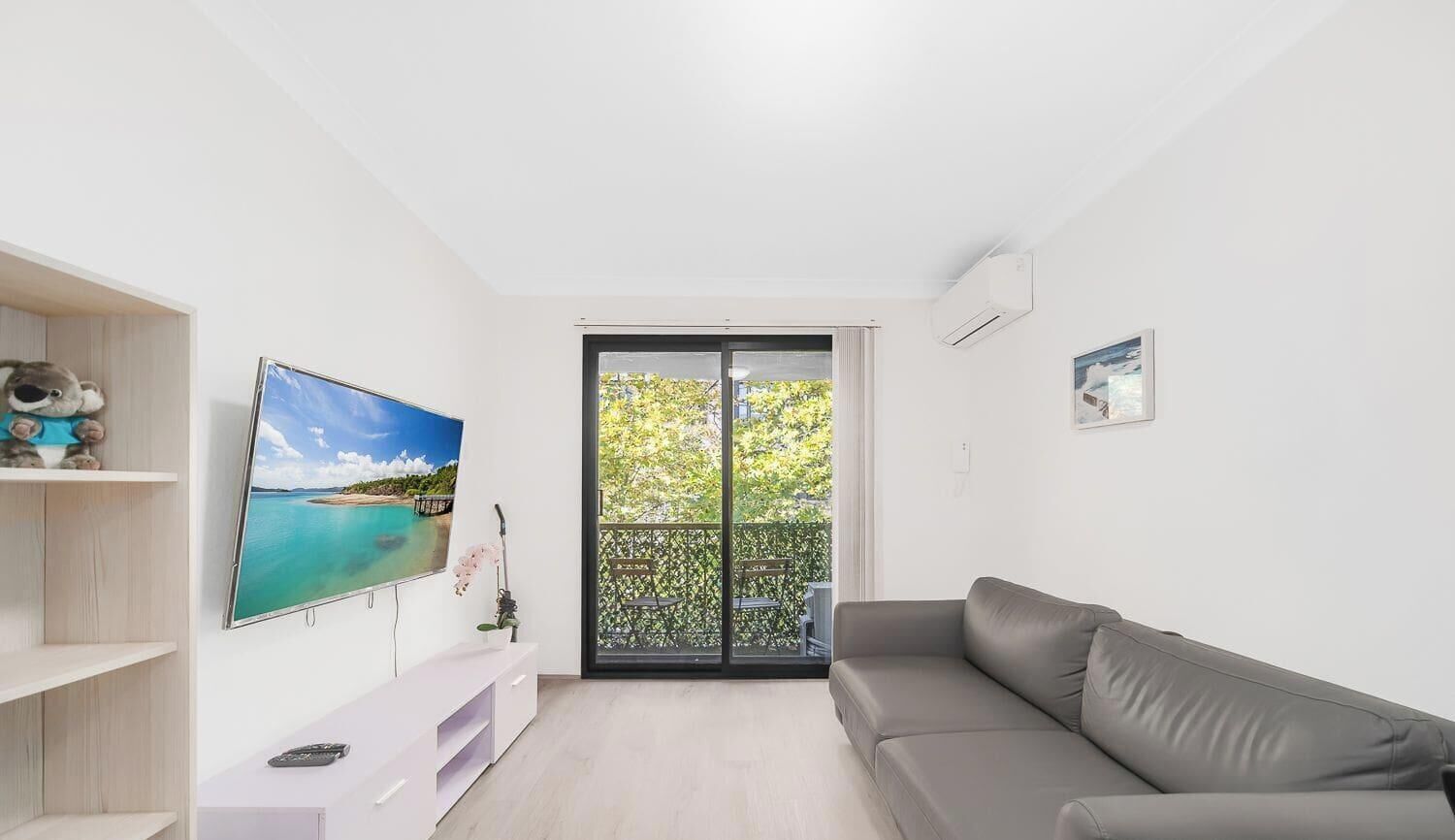 Una sala de estar moderna con un sofá gris, un televisor de pantalla plana montado en la pared y un balcón con vistas a zonas verdes.