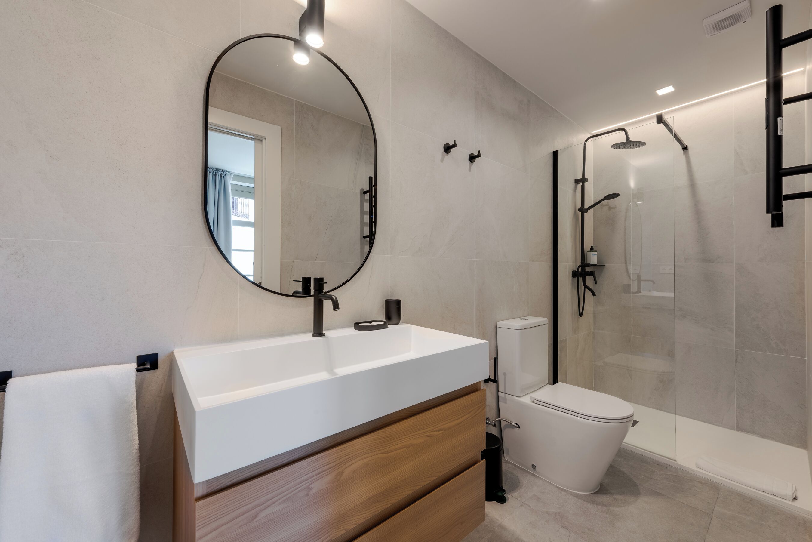 Ein modernes Badezimmer mit einem weißen Waschbecken, einem Holz-Badmöbel, einem runden Spiegel, einer Toilette und einer Dusche mit Glaswand.