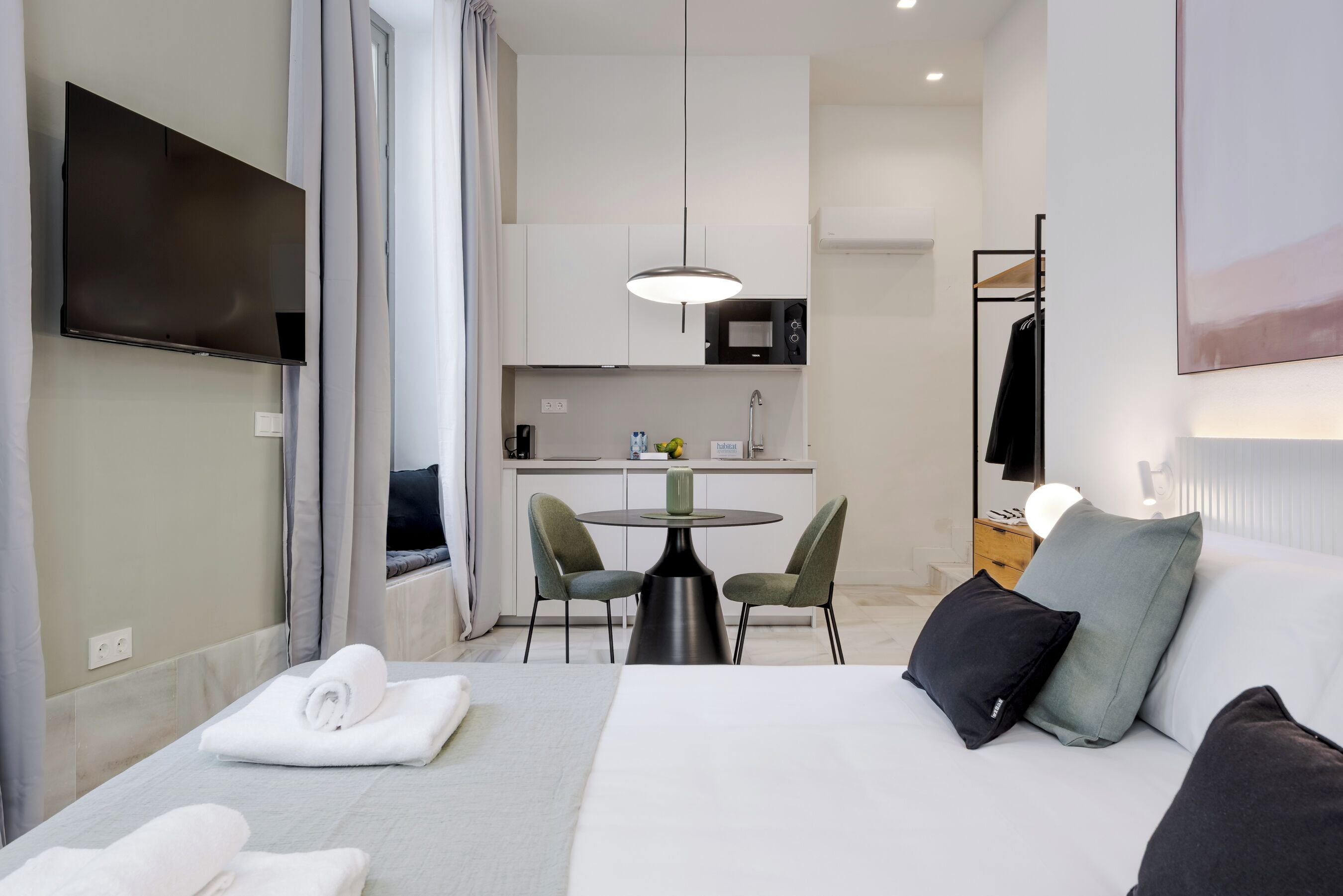 A hotel room with a bed, a TV, a dining table with chairs, a kitchen area, and a coat rack.