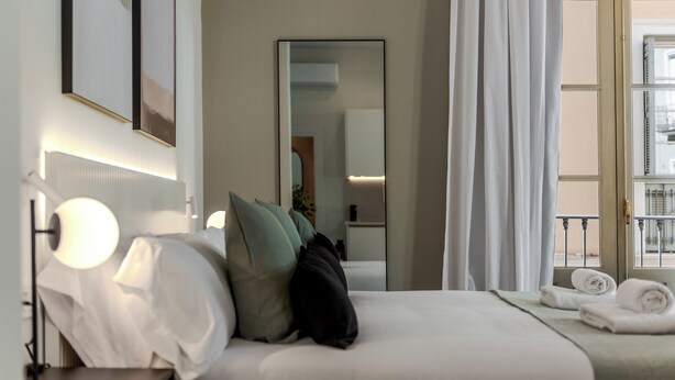 Habitación de hotel con cama, lámparas de noche, mesita de noche, espejo, ventana con cortinas y vistas al exterior.