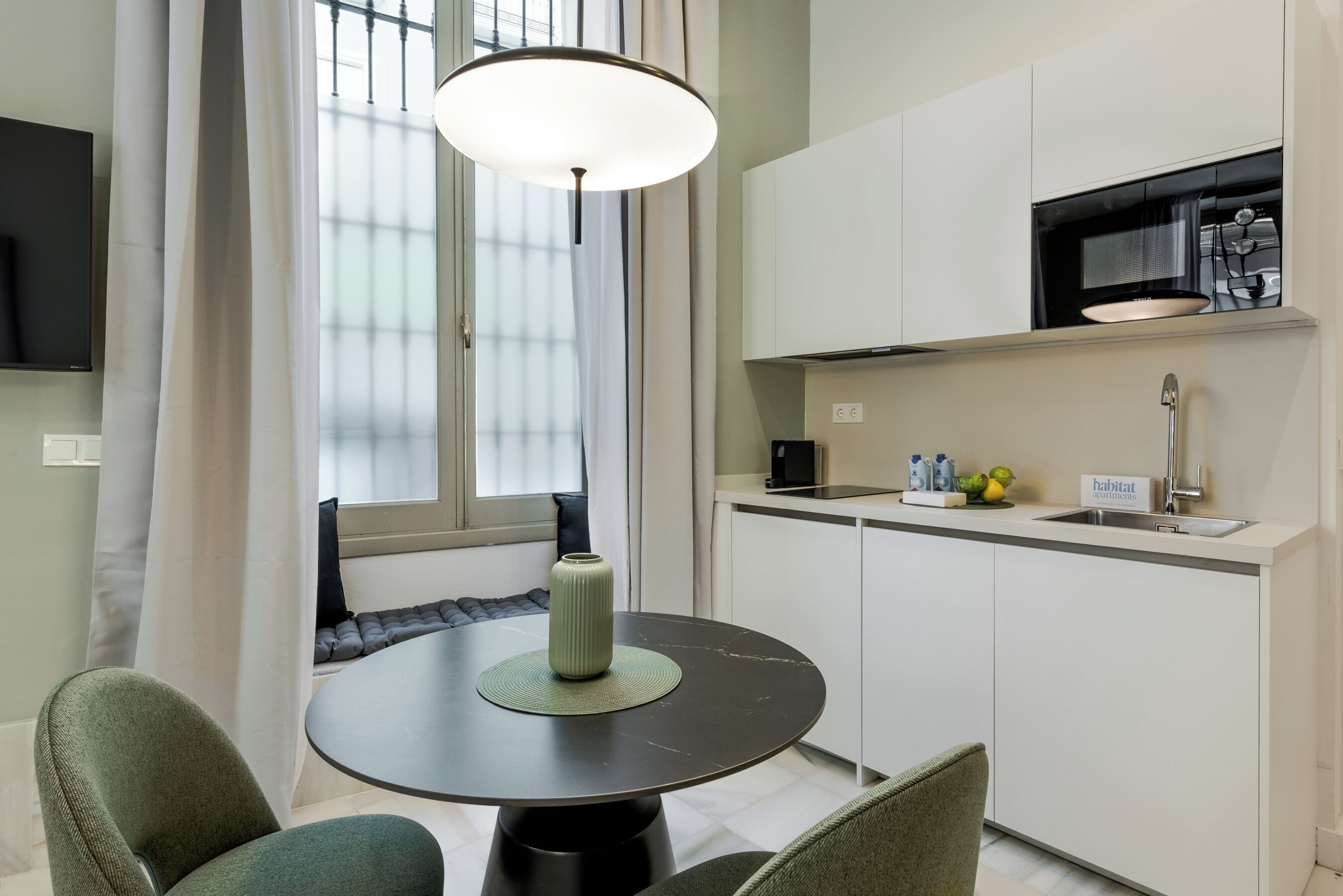 A modern kitchen with a dining area, including a round table with chairs, a hanging light fixture, and a window with curtains.