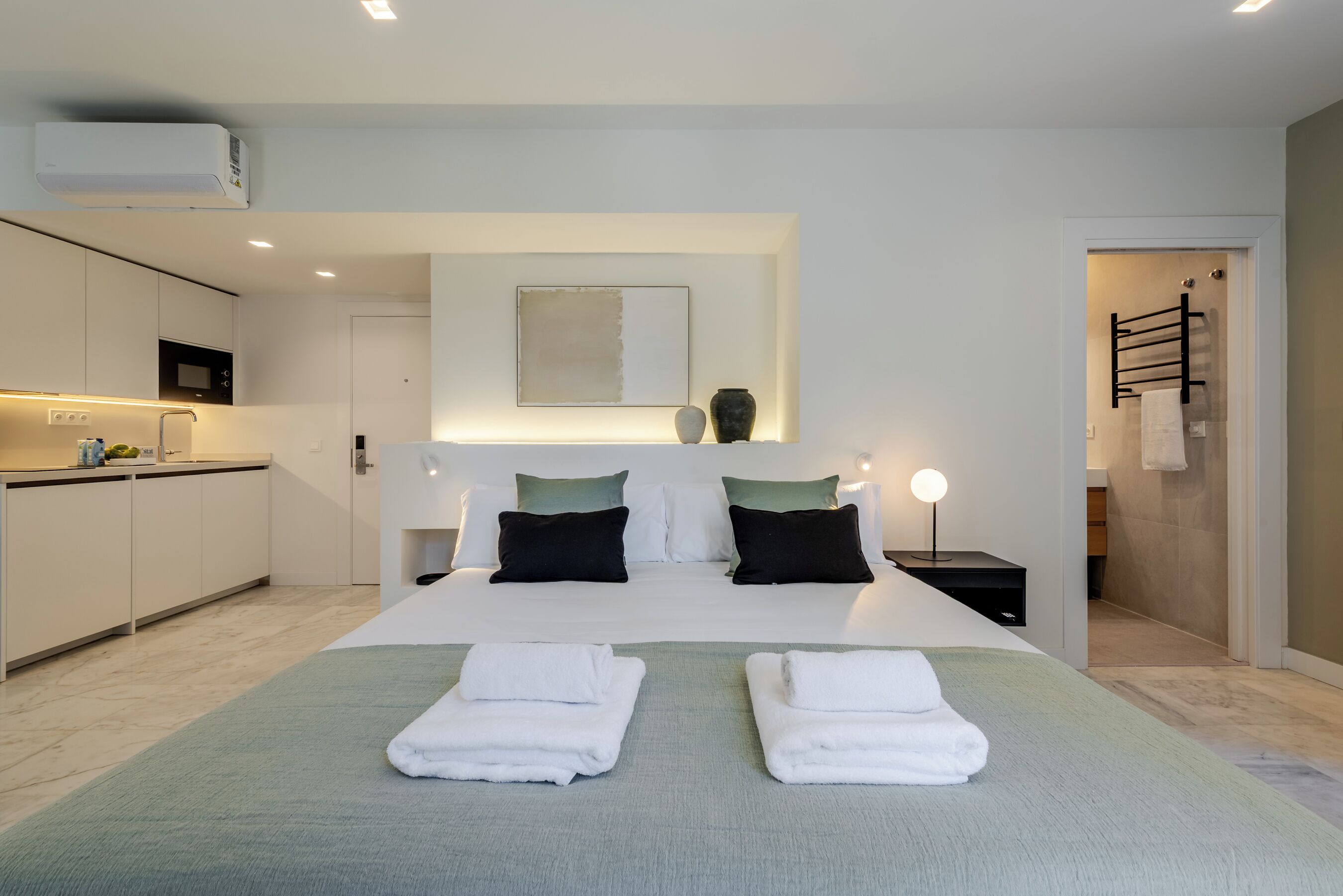 Une chambre d’hôtel moderne dotée d’un grand lit, d’une table de chevet avec une lampe, d’un petit coin cuisine et d’une salle de bain avec un porte-serviettes.