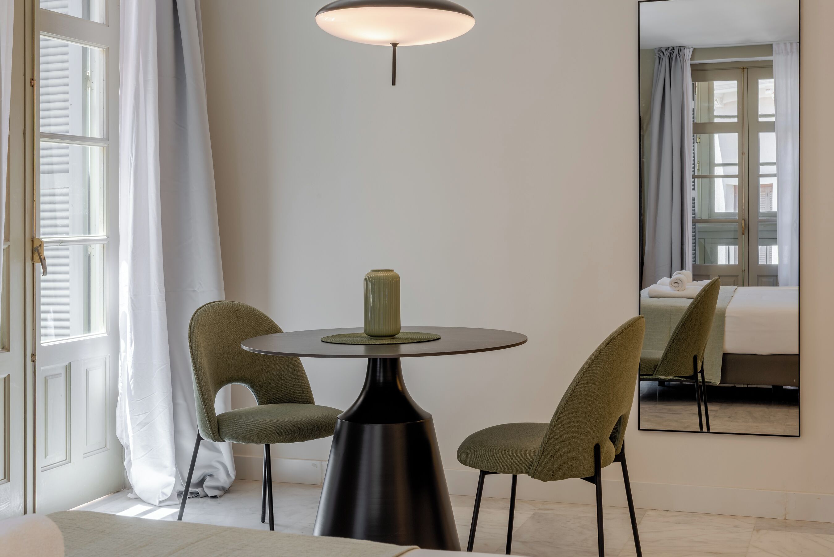 Habitación de hotel con una mesa redonda, dos sillas, un florero, un espejo y una cama.