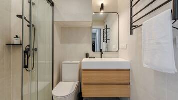 A modern bathroom with a glass shower enclosure, a white toilet, a wooden vanity with a mirror, and a towel rack.