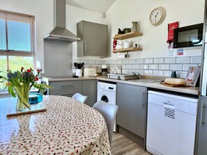 A modern kitchen with a built-in dishwasher, a microwave, and a sink. There is a clock on the wall and a vase with flowers on the table. - The Pepper Pot - Niton, Isle of Wight (Niton)
