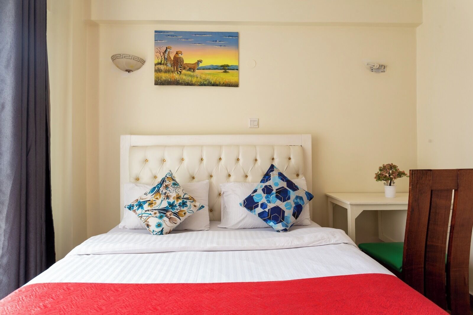 A bed with white bedding, two decorative pillows, a painting of animals on the wall, and a small table with a plant.