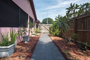 Un chemin pavé mène à une porte en bois, de part et d’autre de laquelle se trouvent des plantes en pot et une clôture en bois.