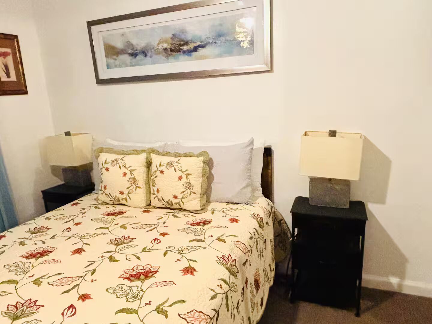 A bedroom with a floral bedspread, two bedside lamps, a framed abstract painting, and a small table with a lamp.