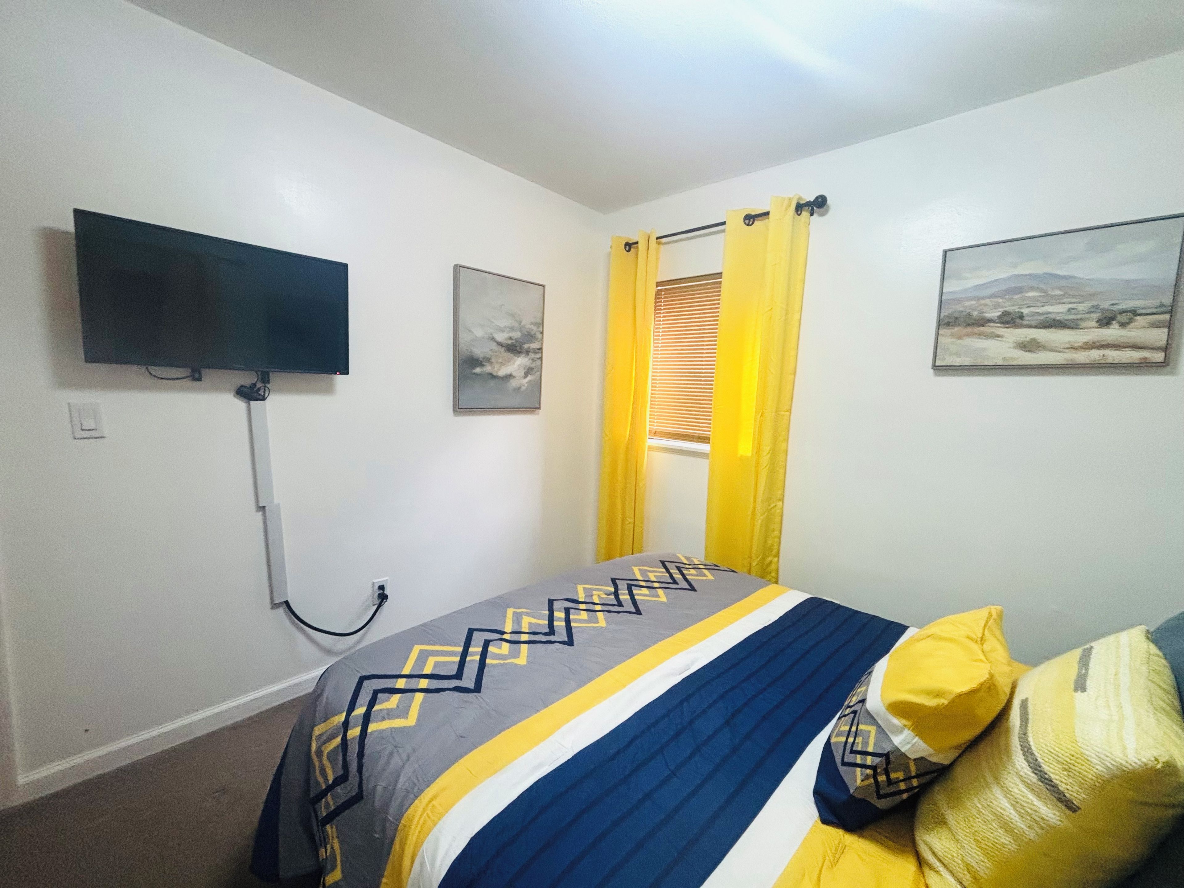 A bedroom with a bed, a flat-screen TV mounted on the wall, a window with yellow curtains, and a landscape painting on the wall.