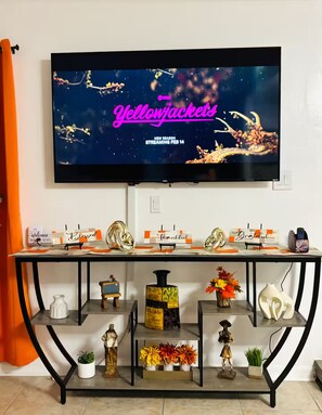 Un téléviseur fixé au mur diffuse un programme intitulé « Yellowjackets », et une date s’affiche : « NOUVELLE SAISON, À PARTIR DU 14 FÉVRIER ». Sous le téléviseur, une table présente divers objets, notamment un panneau « Bienvenue », un panneau « Merci » et des objets décoratifs.