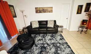 Un salon avec un canapé en cuir noir, des coussins décoratifs à motifs, un tapis noir et blanc, un tableau représentant un paysage urbain encadré accroché au mur, et une petite table avec une lampe.