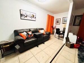 A living room with a black leather sofa, orange curtains, a painting, and a dining area with a chair and table. - Your Tampa Bay Vacation Entertainment Home (Tampa)