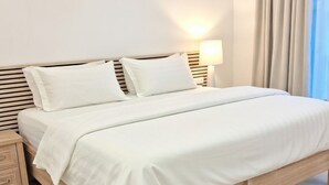 Cama bem arrumada com lençóis brancos em quarto com piso de madeira clara e mesa de cabeceira.