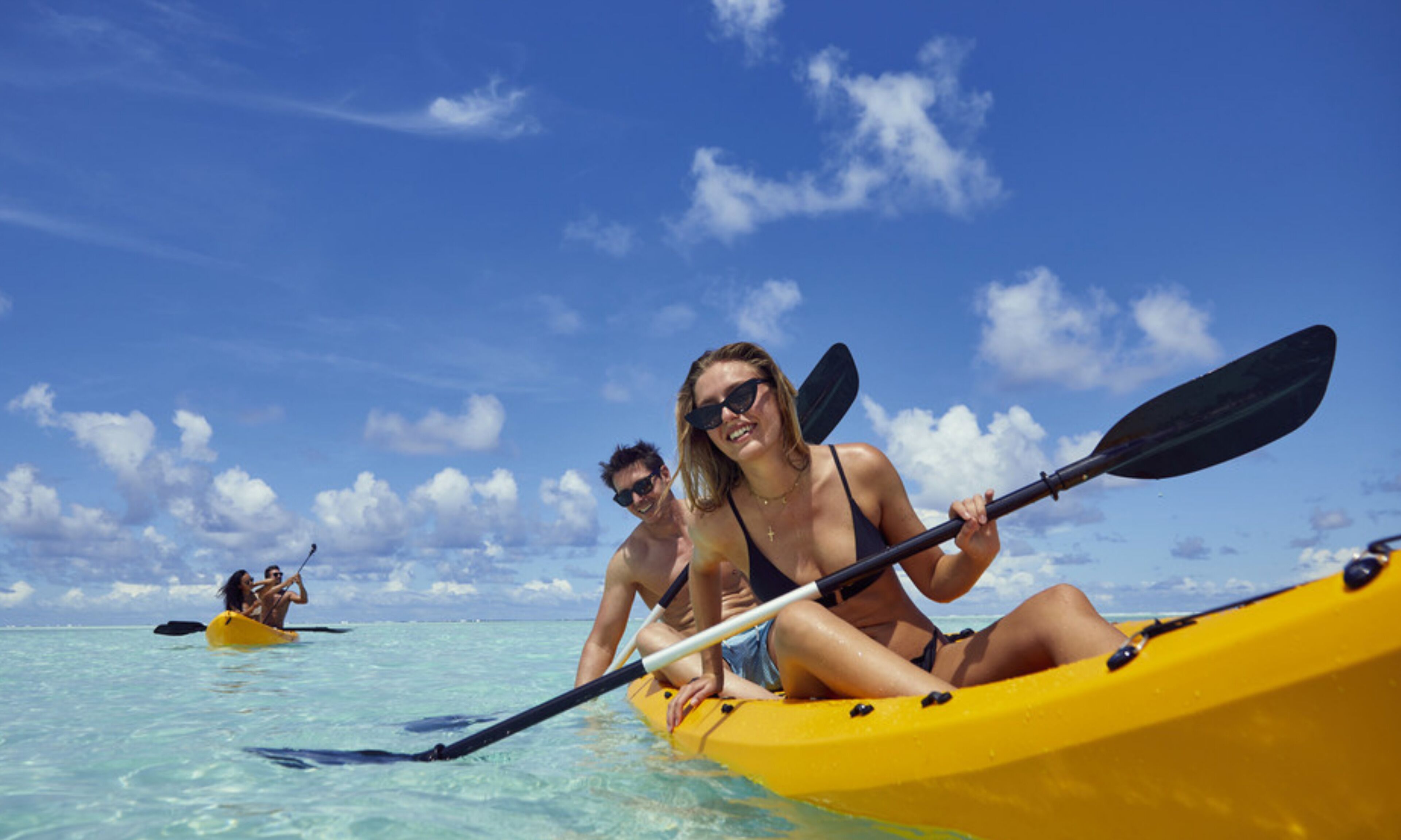 A couple in a yellow kayak on clear turquoise water, with a clear blue sky and distant other kayakers.
