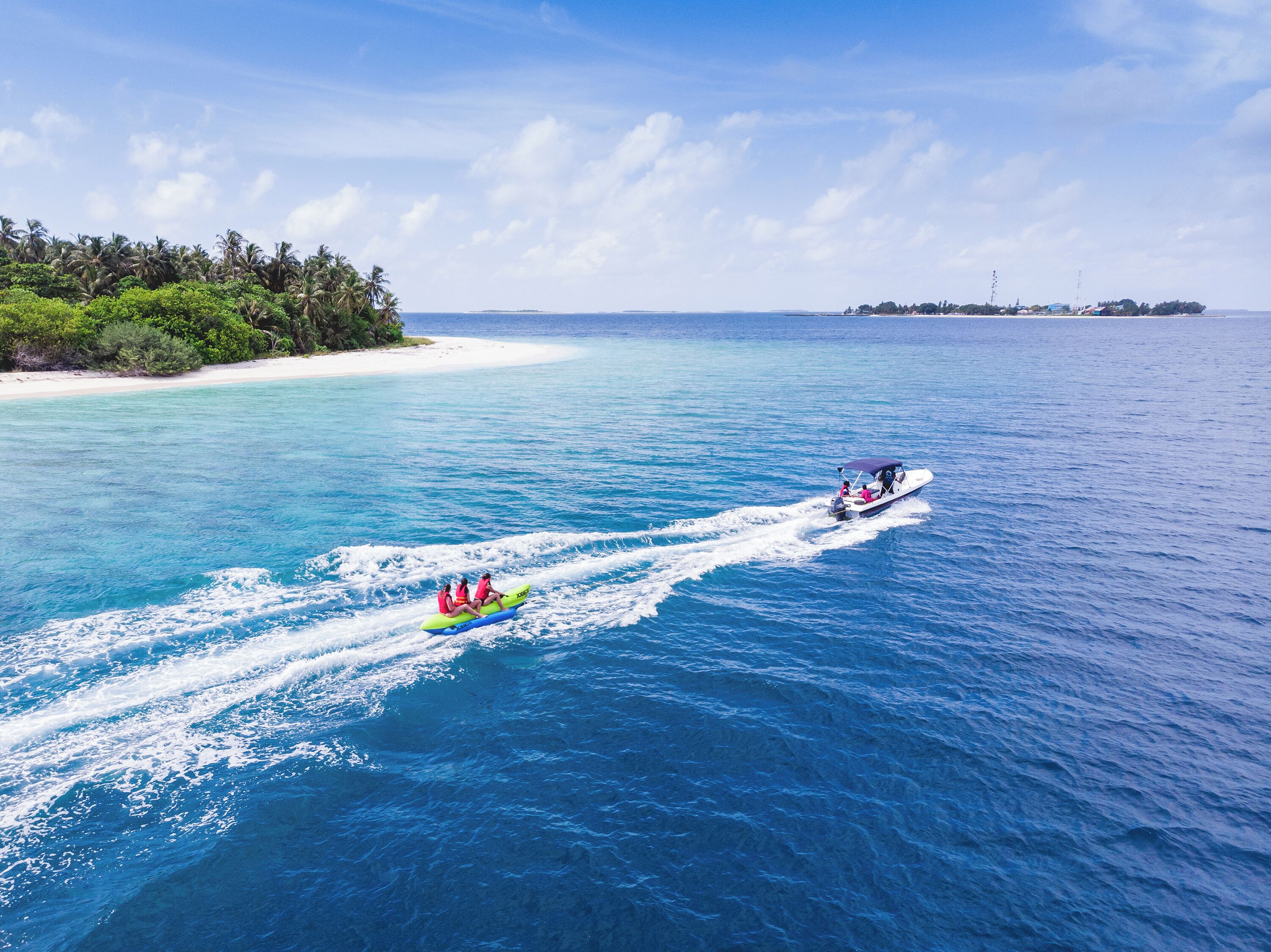 Une mer d’un bleu profond, traversée par un bateau laissant une vague derrière lui, un petit radeau gonflable avec deux personnes à bord, et une côte lointaine parsemée de végétation et de bâtiments.