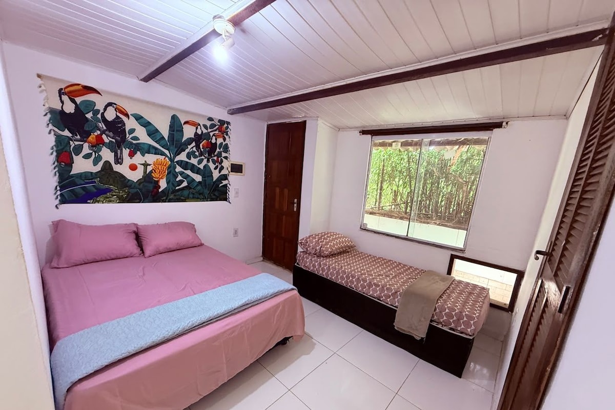A bedroom with a large bed, a smaller bed, a window with a view of greenery, and a wall with a colorful tapestry.
